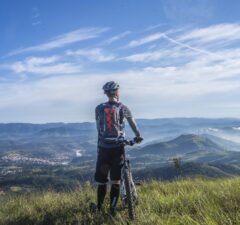 Osoba w stroju kolarskim, stojąca przy rowerze, patrząca w dal na pasmo górskie