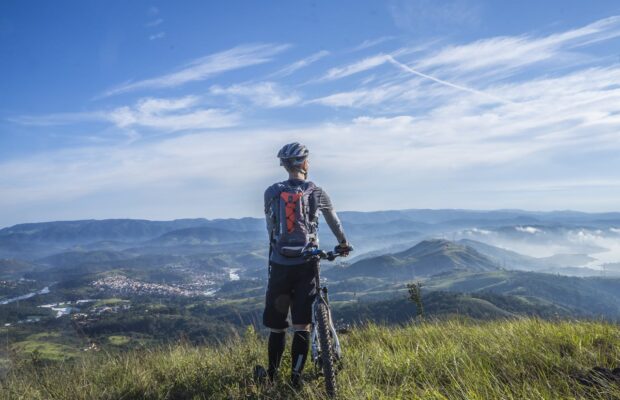 Osoba w stroju kolarskim, stojąca przy rowerze, patrząca w dal na pasmo górskie